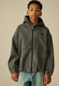 Chaqueta de mezclilla gris con capucha, puños y dobladillo elásticos, que presenta un cierre frontal y dos bolsillos laterales. La prenda se lleva sobre una camiseta morada.