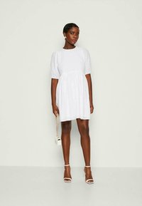 Λευκό φόρεμα με κοντά φουσκωτά μανίκια, υφασμάτινη υφή και φουσκωτό μίντι. Το μοντέλο κρατά μια μικρή λευκή τσάντα και φοράει λευκά πέδιλα με λουριά.