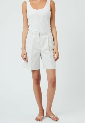 Los pantalones cortos de lino blanco presentan un corte ajustado con pliegues, bolsillos laterales y una cintura alta. Combinados con una camiseta de tirantes blanca ajustada y acanalada.
