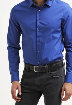 Mężczyzna w dopasowanej, niebieskiej koszuli z długim rękawem, zapinanej na guziki, włożonej do czarnych dżinsów z czarnym paskiem.