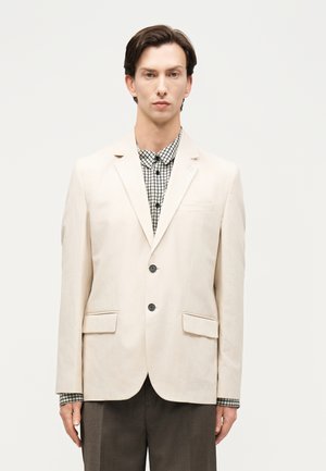 Άνδρας με κοντά σκούρα μαλλιά φοράει εκρού σακάκι πάνω από ασπρόμαυρο πουκάμισο με σχέδια και σκούρο παντελόνι, στέκεται μπροστά σε απλό φόντο.