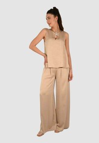 Top beige senza maniche con dettaglio a occhiello e pantaloni abbinati a gamba larga, realizzati in tessuto liscio, caratterizzati da un lieve drappeggio e una vestibilità morbida.