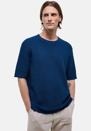 Homme aux cheveux mi-longs portant un pull court bleu marine texturé et un pantalon beige clair, les mains dans les poches.