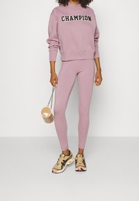 Felpa rosa chiaro con logo "CHAMPION", abbinata a leggings coordinati. L'outfit presenta un design attillato. Scarpe da ginnastica beige e nere indossate.