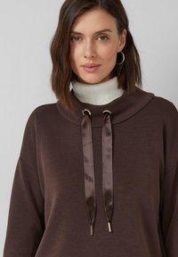 Femme aux cheveux bruns mi-longs portant des boucles d'oreilles créoles, un col roulé blanc côtelé et un haut marron avec des cordons en satin.