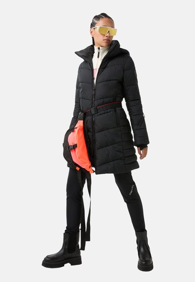 Schwarze Puffermantel mit Kapuze, tailliertem Bund und horizontaler Steppung. Schwarze Leggings und klobige schwarze Stiefel, dazu eine leuchtend orangefarbene Tasche.