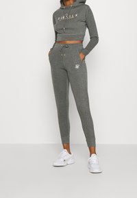 Sudadera gris recortada con letras doradas, combinada con joggers ajustados a juego que cuentan con bolsillos laterales y un logo blanco, usados con zapatillas blancas.