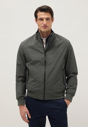 Hombre con chaqueta bomber gris oscuro con cremallera y puños acanalados, y pantalones azul marino, de pie contra un fondo claro y liso.