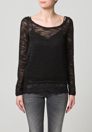 Vrouw in een transparante zwarte lange mouwen top over een zwarte camisole en grijs vervaagde spijkerbroek, staand met handen ontspannen langs de zij.