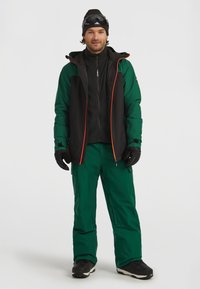Ski outfit med en vandtæt jakke i sort og grøn med orange accenter, parret med grønne isolerede bukser og sorte handsker, sneboots og skibriller.