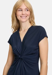 Top enveloppant bleu marine à manches courtes, avec un détail torsadé à la taille et un tissu doux et lisse. Design à col en V.