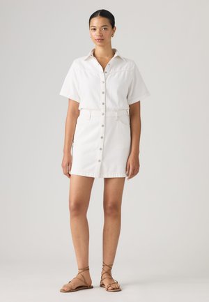Femme debout portant une robe blanche à manches courtes boutonnée avec des poches sur le devant et des sandales plates beiges à brides, sur un fond uni.