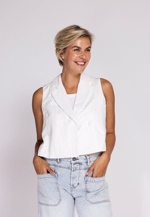 Smiling woman with short blonde hair wears sleeveless white blazer and light-wash jeans, standing with hands in pockets against gray background.