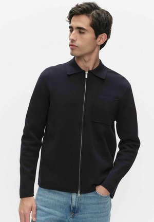 Ung mand iført en marineblå trøje med lynlås og brystlomme samt lyseblå jeans, der ser let til siden mod en ensfarvet baggrund.