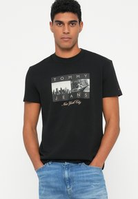 Must musta t-särk mustast puuvillast, millel on graafiline trükk "TOMMY JEANS" ja "New York City" linna silueti ja takso pildiga.