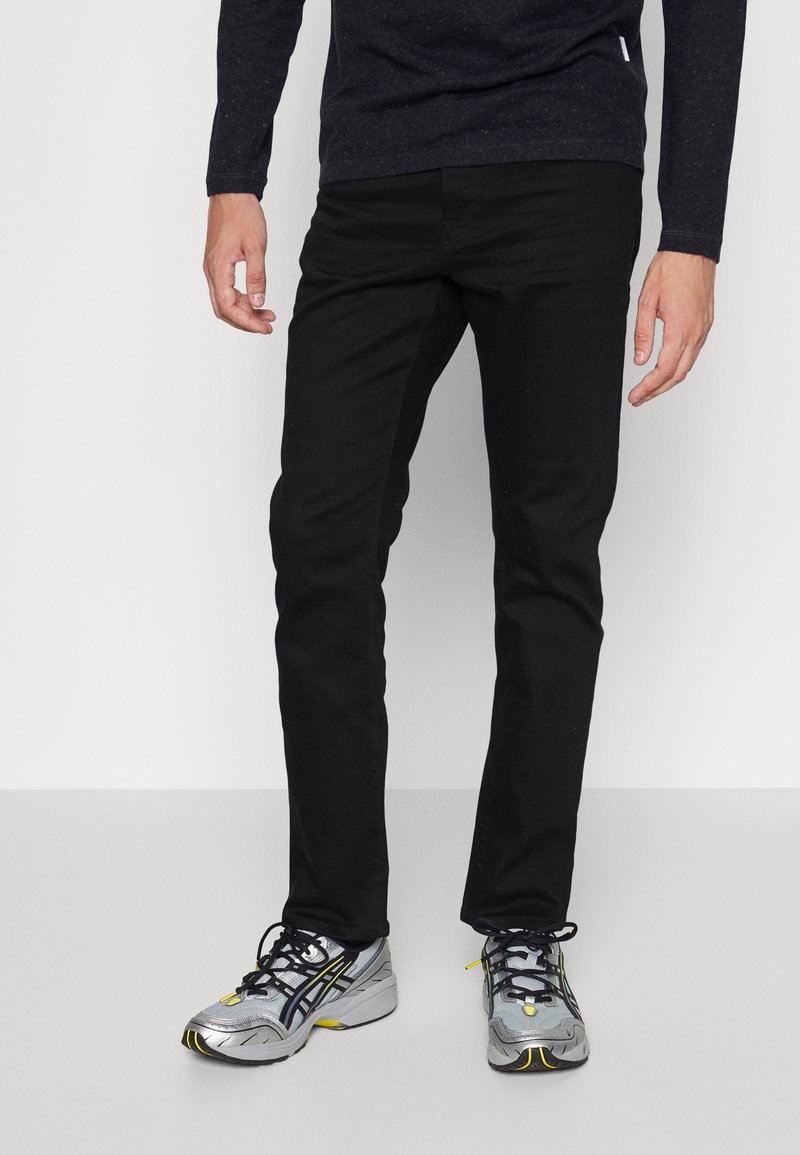 Pantaloni neri slim-fit con una superficie liscia, abbinati a scarpe sportive argento e nere con accenti gialli e una superficie testurizzata.