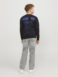 Svart sweatshirt med blå grafisk text p�å ryggen; matchad med ljusgrå jeans och vita sneakers. Kort lockigt hår synligt.