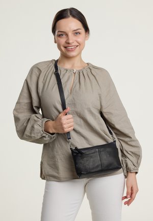 Lächelnde Frau, die eine beige Bluse und weiße Hose trägt und eine schwarze Leder-Umhängetasche vor einem einfarbigen Hintergrund hält.