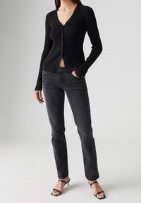 Cárdigan negro con canalé, escote en V y cierre de botones, combinado con jeans rectos gris oscuro y tacones negros con tiras.