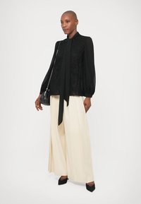 Sieviete, tērpusies melnā garajās piedurknēs blūzē ar kakla sasienamo lentes, plašās krēmkrāsas biksēs, melnās zābakos ar augstu papēdi un melnā pleca somā, stāvot pret balta fona.