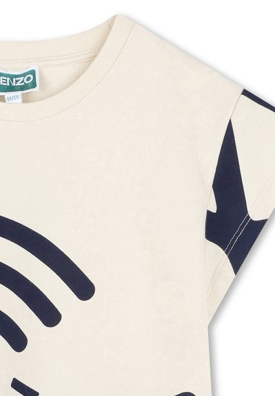 T-shirt en coton couleur crème avec des manches courtes, présentant un motif graphique bleu marine. Étiquette visible près du col. Texture douce, design décontracté.