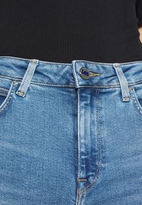 Nahaufnahme von blauen Jeans mit Knopf und Gürtelschlaufen, getragen mit einem schwarzen gerippten Oberteil.