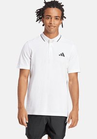 Weißes Poloshirt mit schwarzem Kragen, zwei Knöpfen und schwarzem Adidas-Logo auf der linken Brustseite. Kombiniert mit schwarzen Shorts. Glatte Textur.
