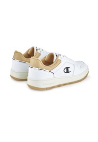 Zapatillas blancas con un acento en color marrón claro, fabricadas en material similar al cuero. Presentan una puntera redonda, un diseño con cordones, y una suela de goma texturizada con el logo de la marca.