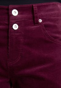 Burgunderfarbene Cordhosen mit sichtbaren Nähten, zwei Metallknöpfen an der Taille und einem kleinen Detail in der Vorderseite der Tasche.