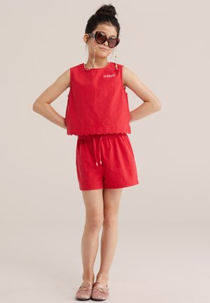 Niña con camiseta sin mangas roja y pantalones cortos con texto "LE SOLEIL", gafas de sol en forma de corazón y zapatos slip-on rosas, de pie con las manos en las caderas.