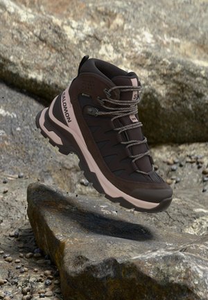 Tmavohnedá a ružová turistická obuv Salomon s hrubou podrážkou a šnúrkami, umiestnená nad kamenistým terénom.