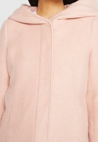 Cappotto cappuccio rosa chiaro in morbido tessuto testurizzato; presenta una linea dritta, chiusura con zip frontale e un leggero dettaglio di cuciture diagonali.