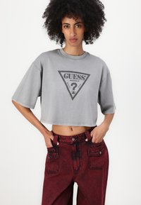 Maglietta corta grigia con logo a triangolo nero. Abbinata a jeans rossi a vita alta con accenti scuri e tasche anteriori.