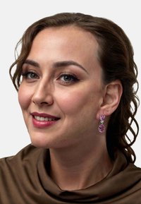 Pink gemstone earrings feature a teardrop shape with silver accents. The model wears a brown fabric drape near the neckline.