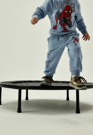 Enfant portant un pantalon de survêtement et un sweat-shirt bleu clair avec un motif Spider-Man, sautant sur un petit trampoline noir. Baskets marron et beige.