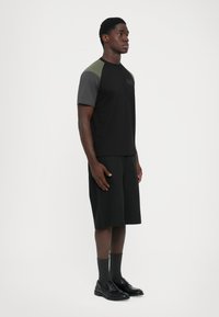Man standing in color-blocked black, gray, and olive t-shirt with "EA7" logo, black shorts, gray socks, and black leather loafers on white background.