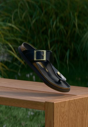 Juoda odinė šlepetė su dideliu auksiniu sagtimi, sklandanti virš medinės suolelio, fone žalios žolės ir augalai.