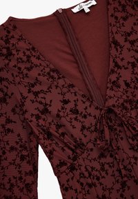 Bordeaux jurk met een diepe V-hals, bloeiende fluwelen patroon, aangesloten taille en strikdetail. Het materiaal heeft een gladde textuur met een lichte glans.