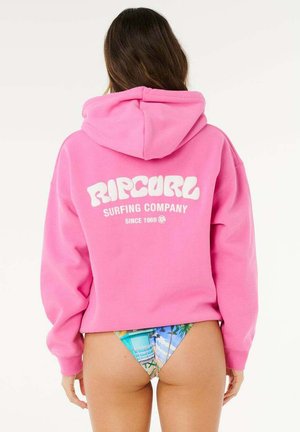 Kobieta ubrana w jaskraworóżową bluzę z kapturem marki Rip Curl oraz kolorowe figi bikini z motywem plażowej wieży ratowniczej, widziana od tyłu.