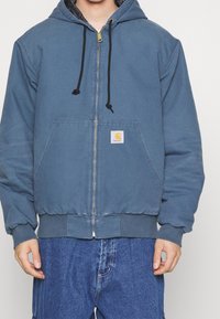 Blauwe zip-up hoodie met een trekkoord capuchon, voorzakken, ribgebreide manchetten en onderrand. Heeft een klein logo patch op de borst.
