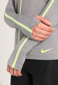 Langarmiges graues Sportshirt mit neon-gelben Streifen, strukturiertem Stoff, Daumenlöchern und Nike-Logo an der unteren Seite.