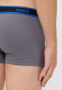 Sive hlače v slogu trunk z oprijetim krojem, ki vključujejo črn pas z modrim napisom. Material ima gladko teksturo.