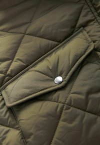 Zeleno jakno iz quiltanega materiala z sijajno površino, ki ima zavihek z žepom, zavarovanim s kovinskim gumbom, ter prikazuje šivano strukturo v obliki diamanta.