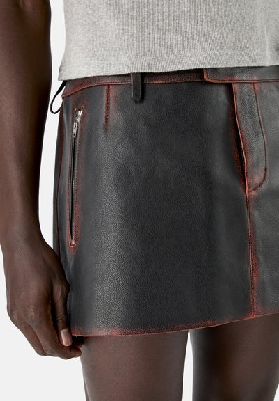 Primo piano di una persona che indossa una gonna in pelle nera con cuciture rosse e una tasca con zip, abbinata a una maglietta grigio chiaro.