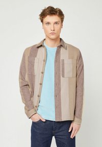 Giacca a camicia a quadri marrone e beige con due tasche sul petto, chiusura a bottoni e una texture morbida, indossata sopra un maglione azzurro chiaro.