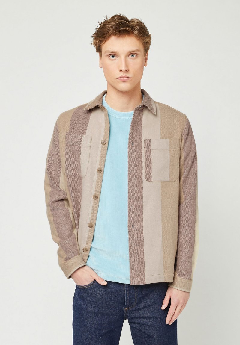 Giacca a camicia a quadri marrone e beige con due tasche sul petto, chiusura a bottoni e una texture morbida, indossata sopra un maglione azzurro chiaro.