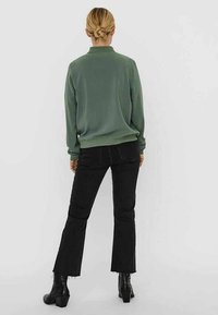 Sudadera verde oliva con escote redondo y puños acanalados, combinada con jeans negros acampanados y botas de tacón negras, vista desde atrás.