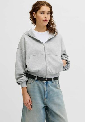 Ung kvinde med bølget hår iført en lysegrå hoodie med lynlås, hvid skjorte, løse blå jeans og sort bælte, stående med den ene hånd i lommen.