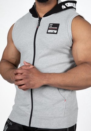 Personne musclée portant un sweat à capuche gris sans manches avec une fermeture éclair, capuche noire et logo "Gorilla Wear", mains jointes devant.