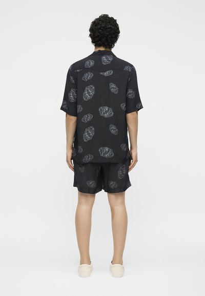 Άνδρας με σγουρά μαύρα μαλλιά στέκεται με την πλάτη, φοράει μαύρο κοντομάνικο πουκάμισο και σορτς με γκρίζα λουλουδάτα σχέδια, λευκά παπούτσια.